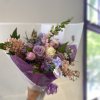 Handheld bouquet of lavender, pink, and cream flowers wrapped in purple paper