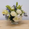 White roses, lilies, and hydrangeas with blue accents arranged in a low clear glass vase