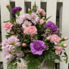 Basket arrangement of pink carnations, lavender daisies, and purple blooms in a white woven basket