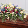 Large mixed flower casket spray with lilies, roses, and blue blooms on a wooden casket