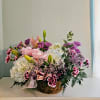 Basket arrangement of white hydrangeas, pink lilies, and purple flowers with a pink ribbon
