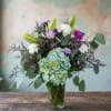 Mixed bouquet with blue hydrangea, purple and white blooms, and lily buds in a clear glass vase.