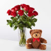 Red roses in a clear vase beside a teddy bear with a red bow