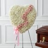 Heart-shaped white floral tribute with pink roses on a stand beside a casket