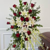 Standing funeral spray with red roses, white lilies, and trailing orchids beside a casket