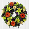 Circular floral wreath with orange gerberas, yellow lilies, cream roses, and purple blooms