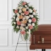 Standing spray of peach roses, pink carnations, and white daisies on an easel beside a casket