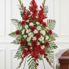 Tall red and white standing spray with carnations and mums on a metal easel beside a wooden casket.