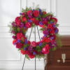 Heart-shaped floral wreath with pink roses and purple flowers on an easel beside a casket