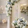 White and pale blue floral wedding arrangements on a draped arch and low pedestal