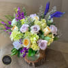 Arrangement of cream and lavender roses with green hydrangeas and purple flowers in a clear vase on a wood slice.