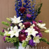 Tall bouquet of white lilies, blue delphinium, and purple blooms in a white vase