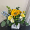 Bright sunflower and yellow roses with blue accents in a white cube vase