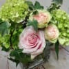 Short arrangement of pale pink roses and green hydrangeas in a clear glass cube vase with trailing ivy