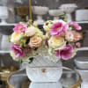 Handbag-shaped white ceramic vase filled with pink anemones, pastel roses, and white hydrangeas on a gold table