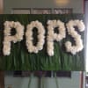 White floral letters spelling "POPS" on a green backdrop.