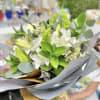 Hand-tied bouquet of white lilies and small white flowers with vivid green accents wrapped in gray and gold paper.