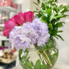 Round glass vase with pink roses, pale lavender hydrangeas, and tall green foliage.