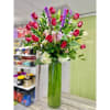 Tall arrangement of red roses, pink lilies, white carnations and purple flowers in a green glass cylinder vase