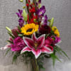 Tall mixed bouquet with pink lilies, sunflowers, purple orchids, and red blooms in a clear glass vase