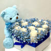Heart-shaped box of white roses with blue filler flowers and a blue teddy bear plush beside it