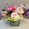Pink and peach roses arranged in a square glass vase with purple accents