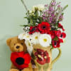 Bouquet in a floral mug beside a teddy bear with red flower accents