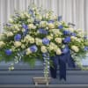 A broad, cascading casket spray in white, blue, and green that reads