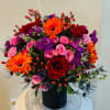 Bright mixed bouquet with red roses, pink spray roses, orange gerberas, and purple blooms in a black vase