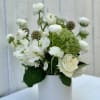 White roses and ranunculus with green accent blooms arranged in a white cylinder vase.