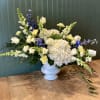 Wide white pedestal vase with white hydrangeas, roses, yellow blooms, and blue delphinium in an asymmetrical design
