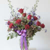 A tall beautiful vase arrangement of red roses, purple delphinium and Queen