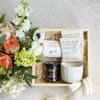 Floral gift box with roses and white blooms beside tea, caramels, fig spread, and a mug