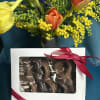 Gift set with yellow and orange tulips in a vase behind a box of chocolate-covered pretzels tied with a red ribbon.