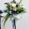 Handheld bouquet of white roses, blue flowers, and fern fronds wrapped in pale blue paper