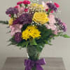 Mixed bouquet of yellow, pink, and purple flowers in a glass vase with a purple ribbon