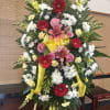 Standing floral wreath with pink lilies, red gerberas, white daisies, and yellow ribbon
