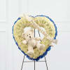 Heart-shaped white flower wreath with a teddy bear and ribbon on a stand
