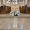 Pastel floral arrangement with a teddy bear on a white pedestal in a church