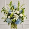 White lilies, roses, blue flowers, and green blooms in a clear glass vase