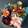 Handheld bouquet of pink, peach, and burgundy flowers wrapped in teal paper