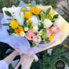Hand-tied bouquet with yellow sunflowers, roses, white lilies, and pastel roses wrapped in yellow and lilac paper.