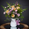 Peach roses and pink flowers in a white pitcher vase with a pink ribbon