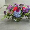 Low glass vase of mixed purple, blue, and red flowers arranged in a wide garden-style centerpiece