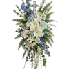 Standing floral spray with white lilies, cream roses, and blue flowers on an easel cross