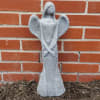 Tall gray stone angel statue with folded arms standing against a red brick wall.