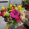 Mixed bouquet of pink gerbera daisies, roses, and yellow tulips in a clear glass vase on a worktable.