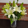 Tall arrangement of white lilies with buds and small white blooms in a glass cylinder vase