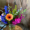 Colorful arrangement with orange gerbera, blue delphinium, and pink stock in a clear glass vase