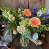 Peach gerbera daisies, cream roses, green hydrangea, and blue delphinium in a clear glass vase